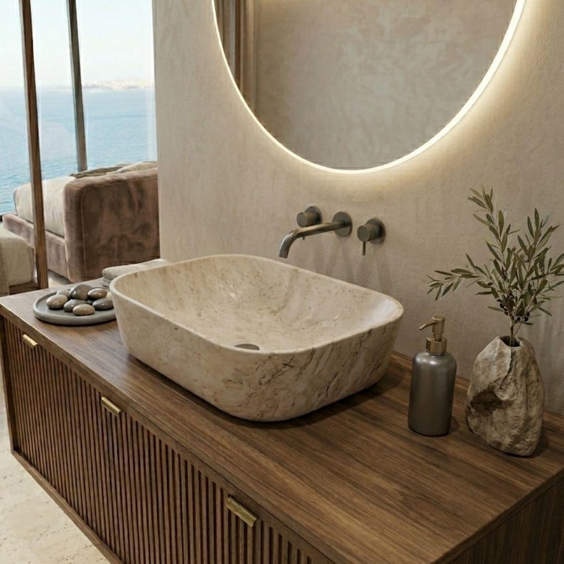 May include: A modern bathroom vanity with a rectangular stone sink. The dark wood cabinet has vertical grooves and gold-toned hardware. A round mirror with a glowing edge hangs above the sink. A soap dispenser and a vase with greenery are on the countertop.