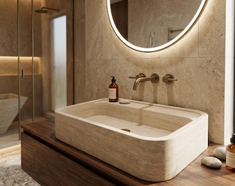 May include: A modern bathroom featuring a rectangular stone sink with a curved front edge, set on a dark wood vanity. A round, illuminated mirror hangs above the sink. A shower with glass doors is visible in the background.
