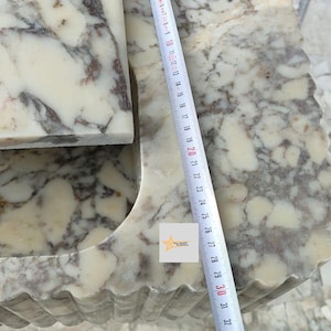 May include: A white and brown marble sink with a fluted exterior design. A white measuring tape is placed vertically across the surface, showing a length of approximately 32 centimeters. A small square logo with a star and text is visible.