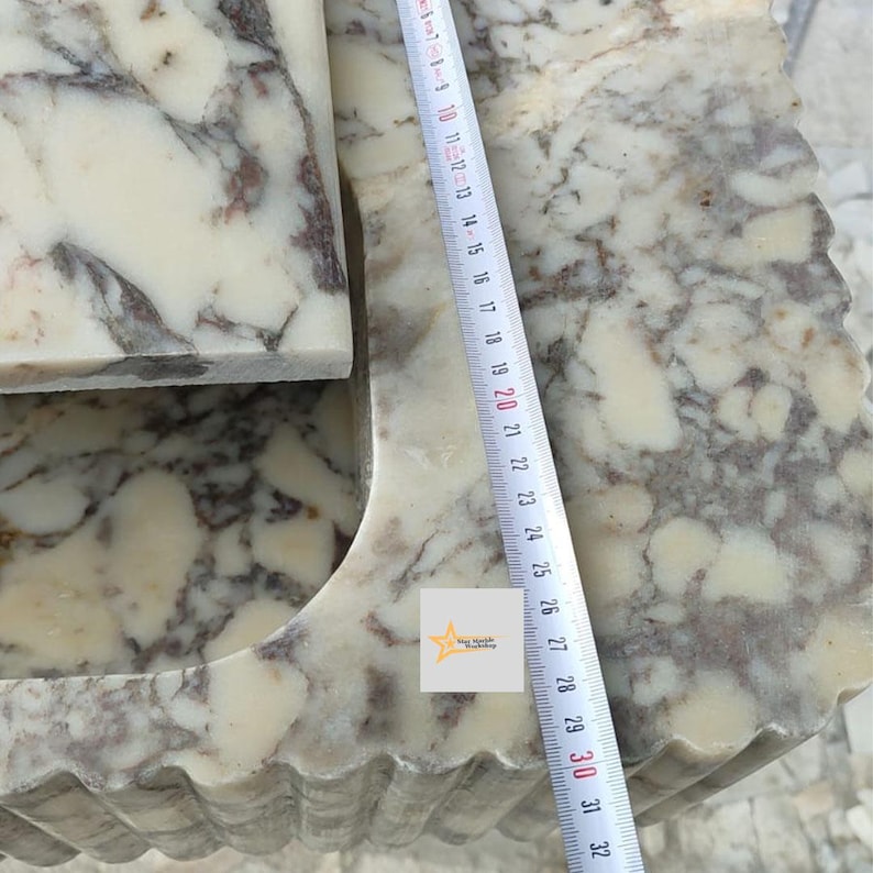 May include: A white and brown marble sink with a fluted exterior design. A white measuring tape is placed vertically across the surface, showing a length of approximately 32 centimeters. A small square logo with a star and text is visible.