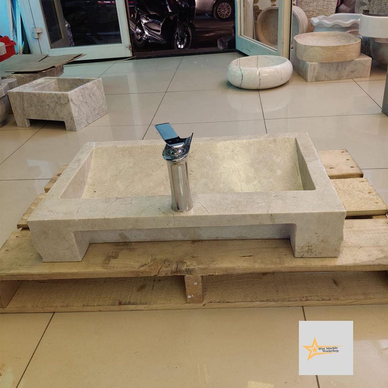 May include: A rectangular stone sink with a chrome faucet, sitting on a wooden pallet. The sink is light beige in color and has a slightly curved design. The faucet is mounted on the top of the sink and has a single handle.