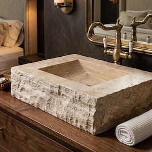 May include: A rectangular vessel sink carved from beige natural stone with a rough, chiseled exterior texture and a smooth interior basin. The sink is positioned on a dark wood surface next to a white rolled towel and a soap dispenser.
