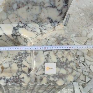 May include: A white and grey marble sink basin with a fluted exterior design. A measuring tape is placed across the top surface, showing a width measurement extending past 95 centimeters. A small square logo with a star is visible on the stone.
