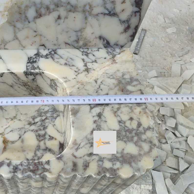 May include: A white and grey marble sink basin with a fluted exterior design. A measuring tape is placed across the top surface, showing a width measurement extending past 95 centimeters. A small square logo with a star is visible on the stone.