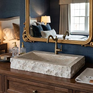 May include: A rectangular stone vessel sink with a rough, natural exterior texture and a smooth interior basin. It features a tall, antique brass faucet with a curved spout and a single lever handle, positioned on a dark wood vanity cabinet.