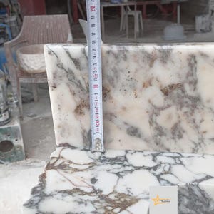 May include: A white marble slab with grey and brown veining, featuring a vertical measuring tape placed against the surface. The tape indicates a height measurement of approximately 20 centimeters from the base to the top edge of the stone piece.