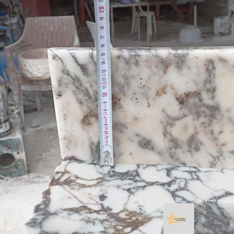 May include: A white marble slab with grey and brown veining, featuring a vertical measuring tape placed against the surface. The tape indicates a height measurement of approximately 20 centimeters from the base to the top edge of the stone piece.