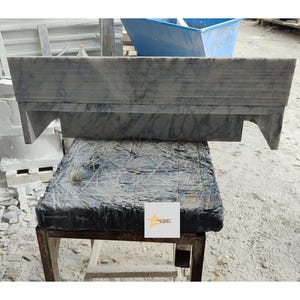 May include: A rectangular grey marble mantelpiece with dark veining and horizontal grooved detailing along the top edge. The piece features two side supports and is resting on a dark, textured base with a small white square logo featuring a yellow star.