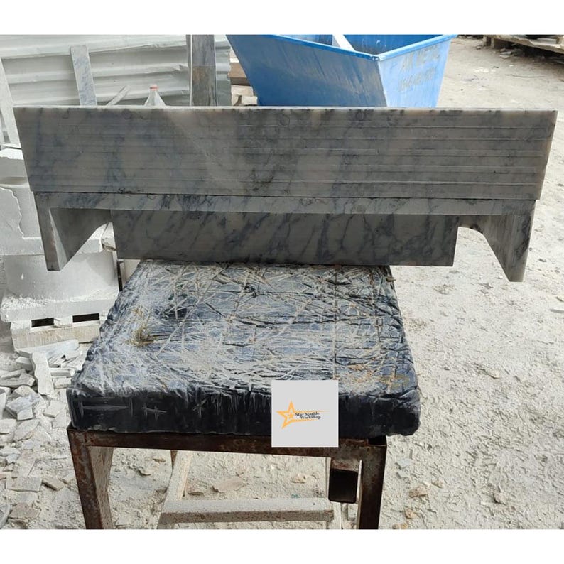 May include: A rectangular grey marble mantelpiece with dark veining and horizontal grooved detailing along the top edge. The piece features two side supports and is resting on a dark, textured base with a small white square logo featuring a yellow star.