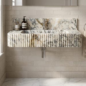 May include: A wall-mounted marble sink featuring a fluted exterior design with grey and gold veining. The basin includes a chrome faucet and a matching marble backsplash. A dark brown soap bottle and a small cup are placed on the left side.
