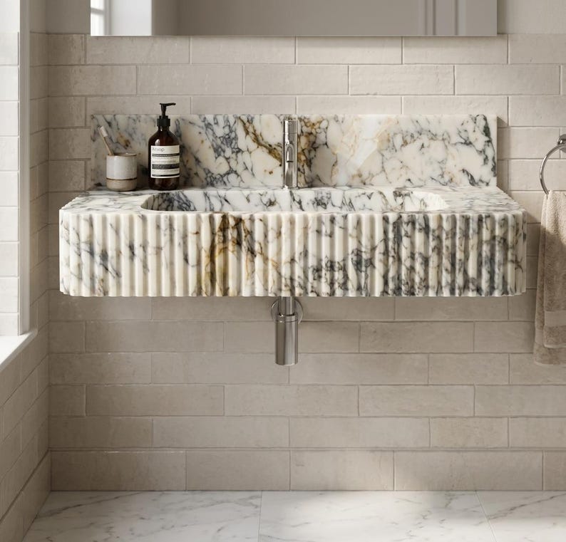 May include: A wall-mounted marble sink featuring a fluted exterior design with grey and gold veining. The basin includes a chrome faucet and a matching marble backsplash. A dark brown soap bottle and a small cup are placed on the left side.