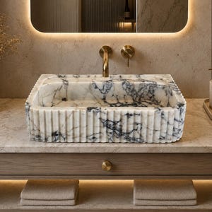 May include: A rectangular bathroom sink carved from white marble with prominent dark grey veining. The exterior features a vertical fluted texture. The basin is positioned on a stone countertop with a brass faucet and a single handle control.