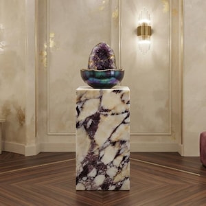 May include: A large, polished purple amethyst geode sits atop a dark, iridescent bowl, which is placed on a tall, rectangular pedestal made of white and purple veined marble. The display is centered in a room with matching marble wall panels.