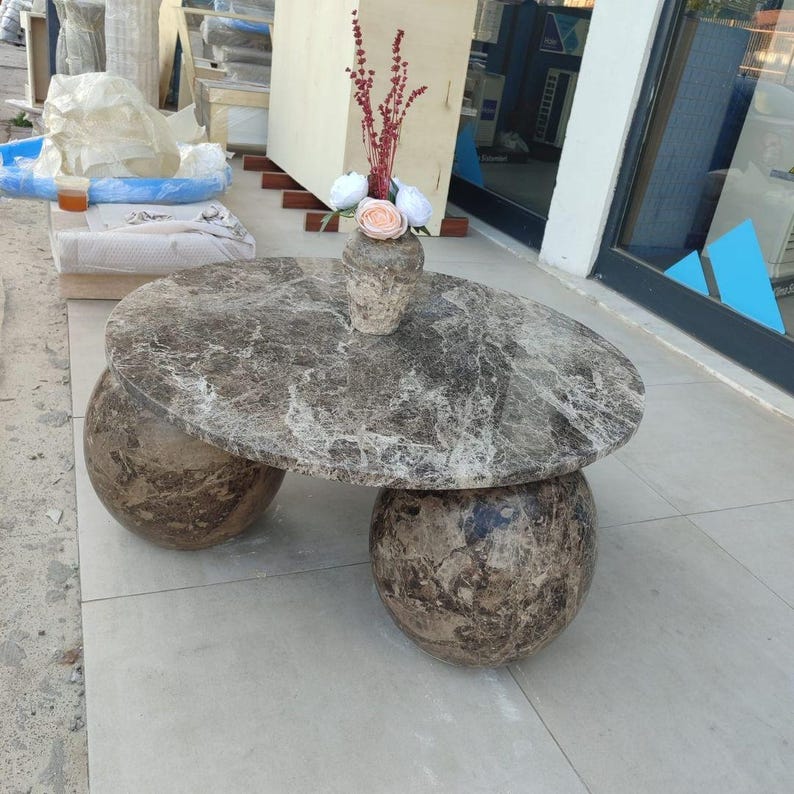 Op de afbeelding: Ovale marmeren salontafel met twee bolvormige bases. De tafel heeft een grijs-bruin gemarmerd patroon. Bovenop staat een kleine vaas met bloemen. De tafel is gemaakt van steen en is een uniek meubelstuk.