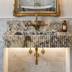 May include: A wall-mounted marble sink featuring a fluted front design with dark grey veining. The sink includes a brass faucet, matching brass plumbing fixtures, and a small soap dish. A gold-framed mirror hangs above the sink.