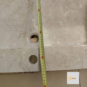 May include: A close-up of a light gray marble sink with two holes. A measuring tape is placed along the side of the sink, showing a measurement of 49 inches.