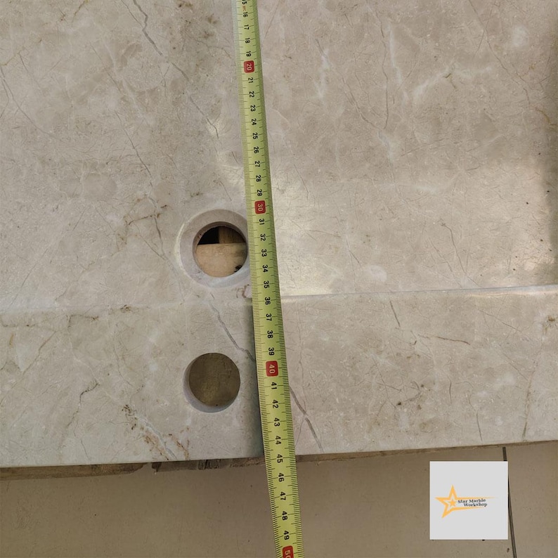 May include: A close-up of a light gray marble sink with two holes. A measuring tape is placed along the side of the sink, showing a measurement of 49 inches.