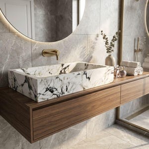 May include: A rectangular marble sink with black veining sits atop a wooden vanity with two drawers. A gold faucet is mounted on the wall above the sink. A round mirror with a light is above the sink. The bathroom decor includes a vase, perfume bottles, and rolled towels.