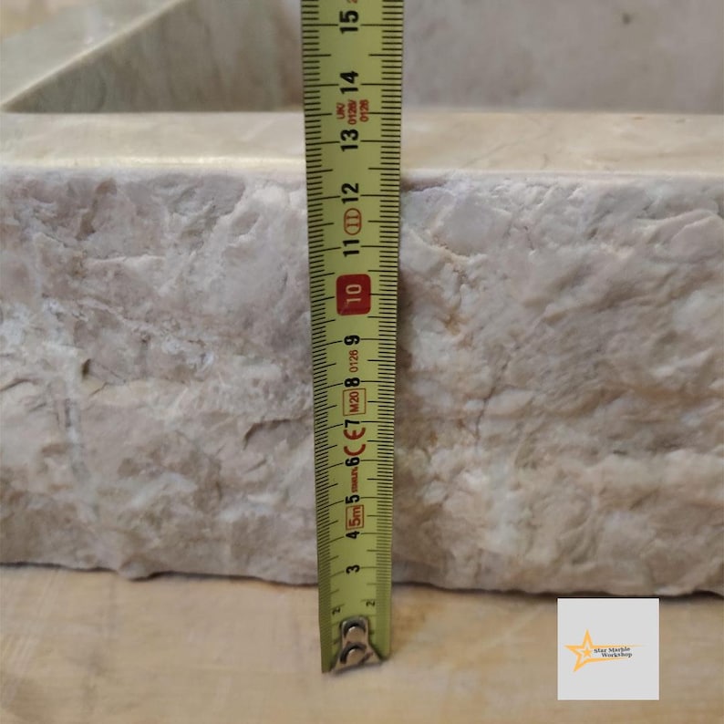 May include: Close-up of a white marble sink with a yellow measuring tape next to it. The tape measures 10 inches.