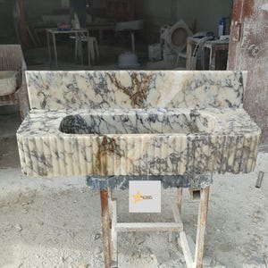 May include: A rectangular marble sink featuring a cream and brown veined pattern with a fluted exterior design. The sink includes a tall matching backsplash and a central basin. A small square label with a yellow star logo is attached to the front.