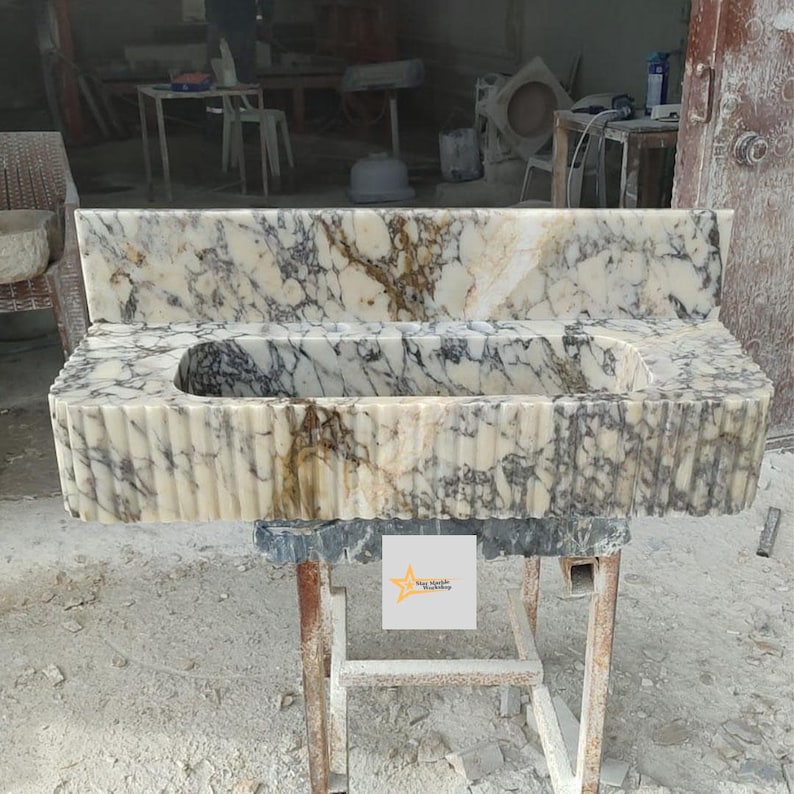 May include: A rectangular marble sink featuring a cream and brown veined pattern with a fluted exterior design. The sink includes a tall matching backsplash and a central basin. A small square label with a yellow star logo is attached to the front.