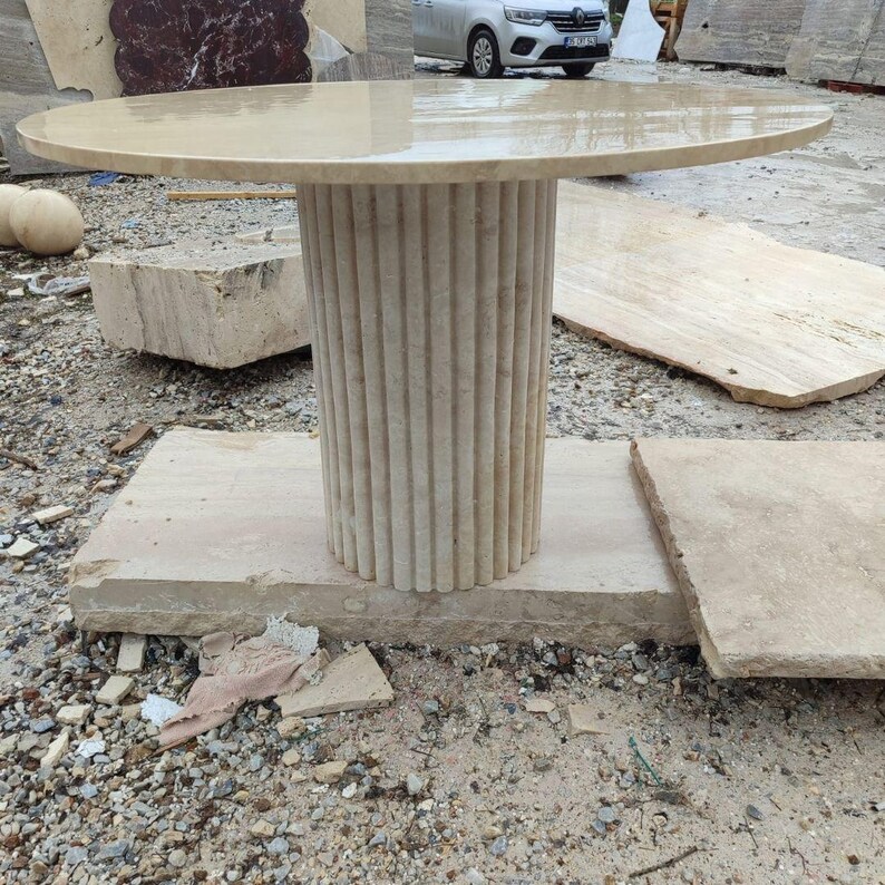 May include: A beige stone table with a round top and a fluted column base. The table is set on a rectangular stone base. The table's top is approximately 48 inches in diameter.