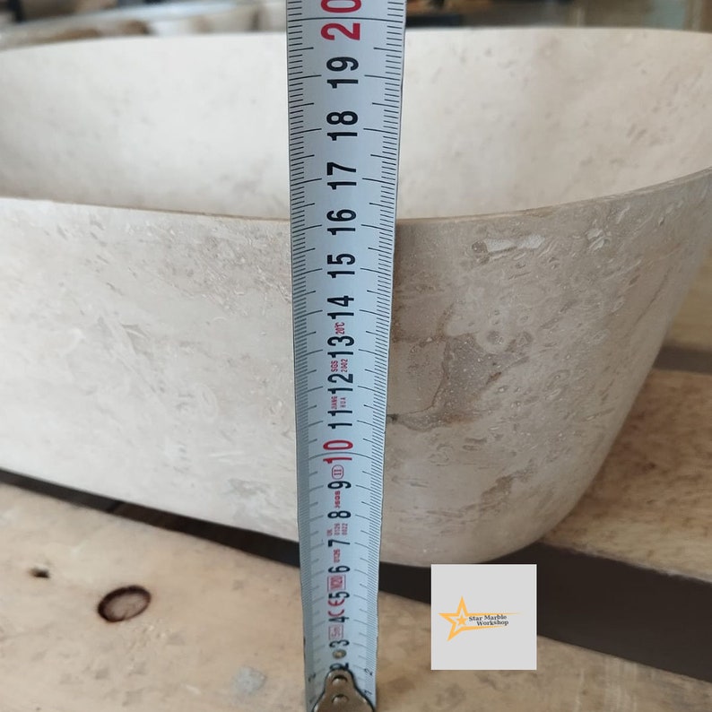 May include: A beige rectangular stone sink with rounded corners. A measuring tape shows the sink's depth. The sink's depth is approximately 10 inches. The sink is made of a light-colored stone with natural variations in color and texture.
