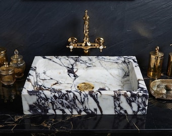 May include: A rectangular bathroom sink crafted from white marble with prominent dark purple and black veining. The basin features a polished finish and includes a circular gold-toned drain cover with an intricate filigree pattern.