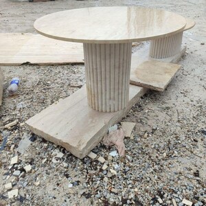 May include: A light beige stone table with a round top and a fluted column base. The table sits on a rectangular stone base. The table is approximately 36 inches in diameter.