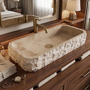 May include: A rectangular stone vessel sink with a rough, chiseled exterior and a smooth interior basin. The sink features a gold-toned drain and is paired with a matching gold-toned faucet, set upon a dark wood vanity with gold hardware.