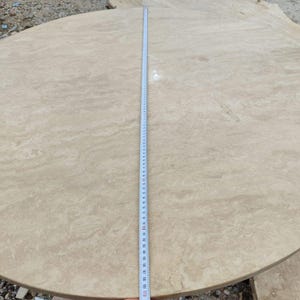 May include: A large, round, beige stone table top with a ruler laid across the center. The ruler measures up to 110 inches. The stone has a natural, marbled appearance with subtle variations in color and texture. The table top is unfinished.