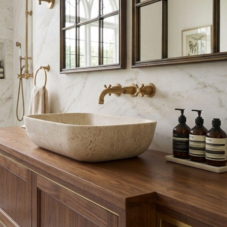 May include: A bathroom vanity with a rectangular stone sink, brass fixtures, and a dark wood cabinet. The countertop holds three brown bottles with black labels. The background features a marble wall and a window.