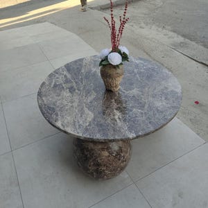 Op de afbeelding: Een ronde marmeren salontafel met een donkergrijs en bruin gemarmerd patroon. De tafel heeft een bolvormige voet en een decoratieve vaas met witte bloemen en rode stelen.