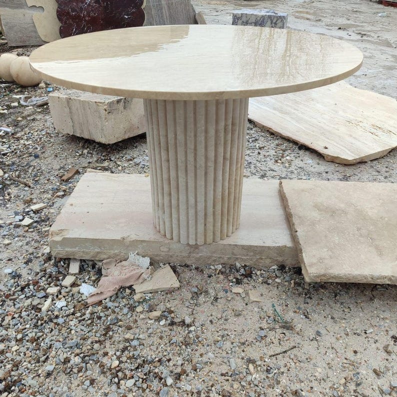 May include: A beige stone table with a round top and a fluted column base. The table is set on a rectangular stone base. The table's top is approximately 36 inches in diameter.