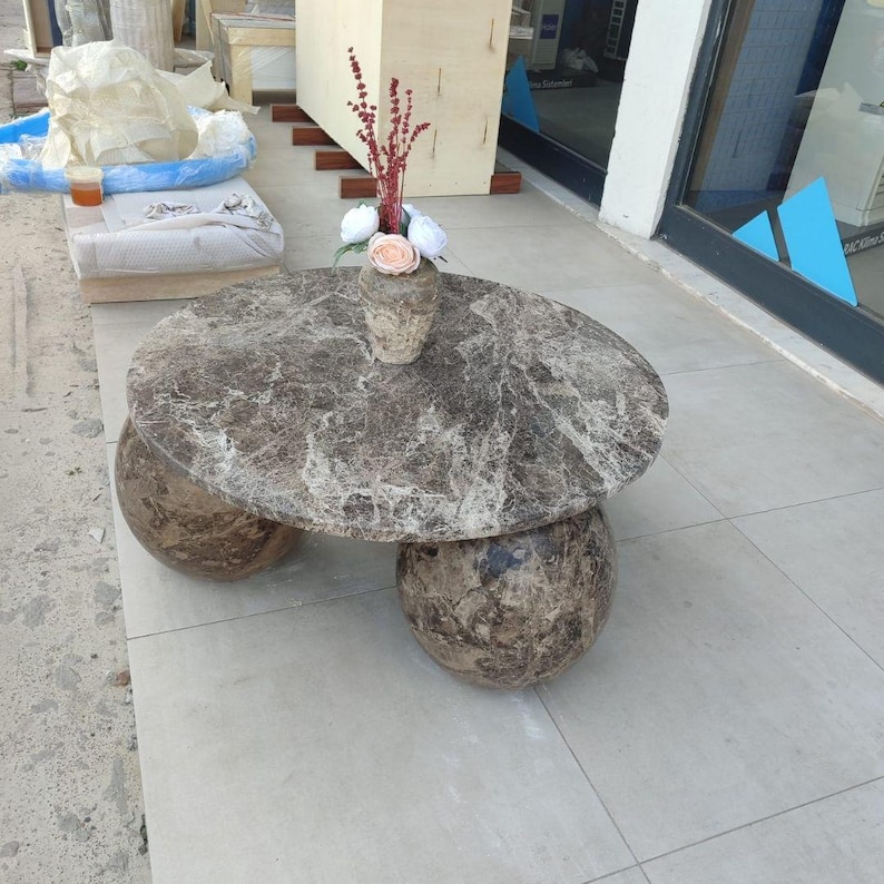 Op de afbeelding: Een ronde marmeren salontafel met een bruin en grijs gemarmerd patroon. De tafel heeft een cirkelvormig blad dat wordt ondersteund door drie bolvormige poten. Een kleine vaas met bloemen staat op het tafeloppervlak.