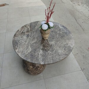 Op de afbeelding: Een ronde marmeren salontafel met een grijs en bruin patroon. De tafel heeft een enkele, afgeronde voet. Een vaas met witte bloemen en rode stelen staat bovenop de tafel.