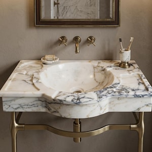 May include: A white marble bathroom vanity sink with grey veining, featuring a built-in soap dish and toothbrush holder. The sink is supported by a brass metal frame and includes matching brass wall-mounted faucet handles and a central spout.