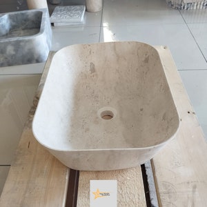May include: A rectangular, beige stone sink with rounded corners and a central drain hole. The sink is set on a wooden surface. The Star Marble Workshop logo is visible in the bottom right corner.