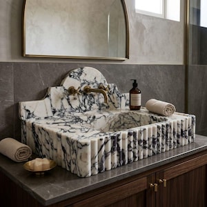 May include: A marble bathroom sink with a black and white veined pattern. The sink has a fluted edge design and brass fixtures. A rolled beige towel and soap dish are on the countertop. A brown soap dispenser and a gold-framed mirror are in the background.