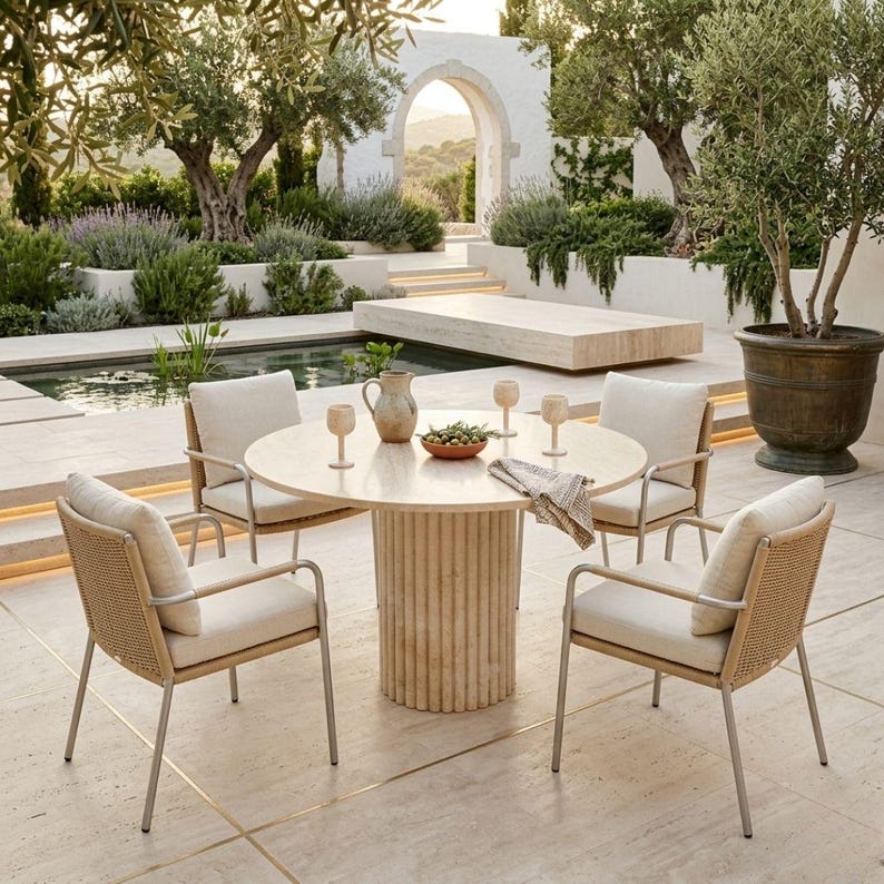 May include: Outdoor dining set featuring a round table with a light-colored stone base and top, surrounded by four chairs with woven backs and cream-colored cushions. The scene is set in a garden with a pool and olive trees.