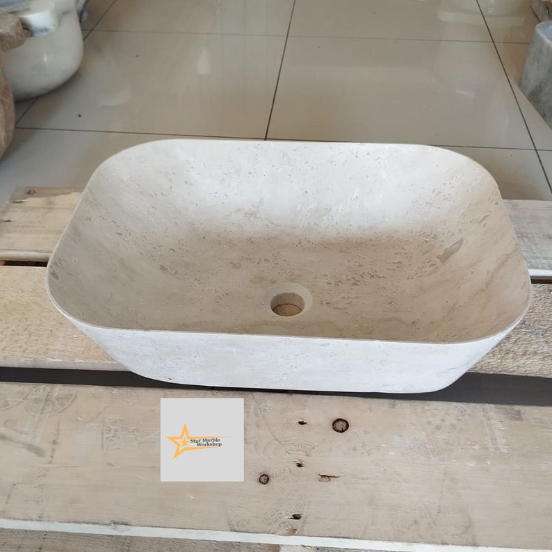 May include: A rectangular, beige stone vessel sink with a rounded interior and a central drain hole. The sink is made of a light-colored stone with natural veining. The sink is displayed on a wooden pallet.