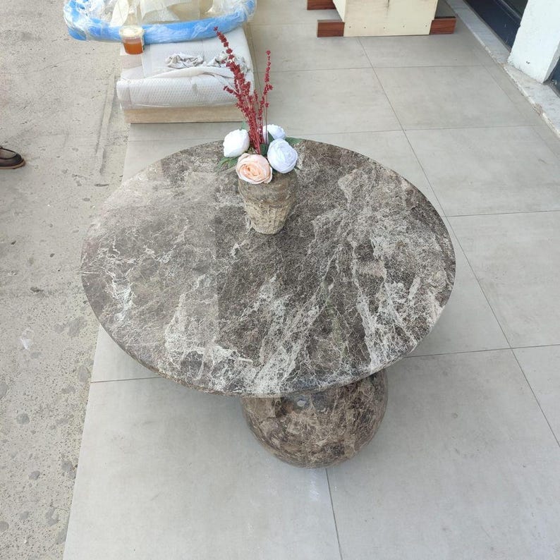 Op de afbeelding: Een ronde marmeren salontafel met een grijs en wit patroon. De tafel heeft een cirkelvormig blad en een bolvormige voet. Een kleine vaas met bloemen staat op de tafel.