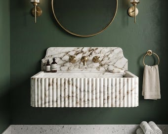 May include: A wall-mounted white marble sink with a fluted front design and a matching marble backsplash. The sink features brass hardware, including a central faucet and two cross-handle valves, set against a dark green wall with a round mirror.