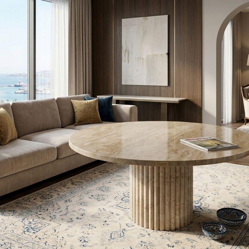May include: A beige sectional sofa with decorative pillows, a round marble coffee table, and a patterned rug. The coffee table has a fluted base and a glossy top. A magazine rests on the table.