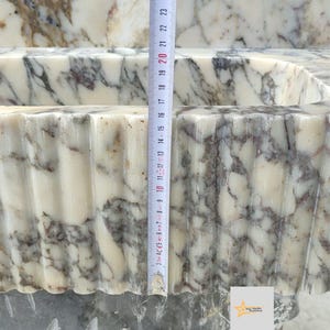 May include: A white marble sink with grey and brown veining, featuring a fluted exterior design. A vertical measuring tape is placed against the front, showing a height measurement of approximately 23 centimeters from the base to the top rim.