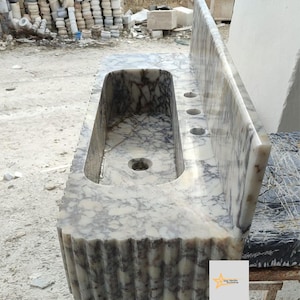 May include: A rectangular marble sink featuring a cream and dark grey veined pattern. The basin has a central drain hole and three faucet holes on the flat deck. The front and side edges of the sink display a vertical fluted, ribbed texture.