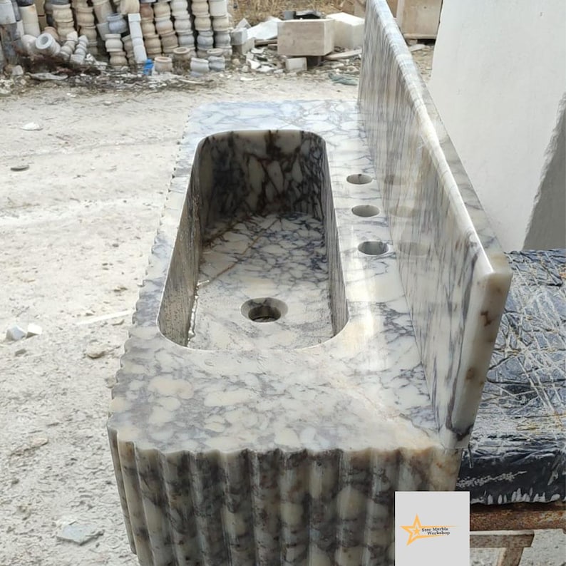 May include: A rectangular marble sink featuring a cream and dark grey veined pattern. The basin has a central drain hole and three faucet holes on the flat deck. The front and side edges of the sink display a vertical fluted, ribbed texture.