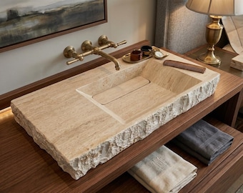May include: A rectangular beige travertine stone sink with a rough, chiseled edge and a smooth basin featuring a central drain cover. A wall-mounted brass faucet with two handles is positioned above the sink, accompanied by a small tray and comb.