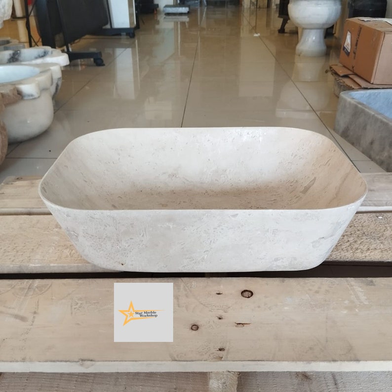 May include: A rectangular, light-colored stone sink with rounded corners. The sink is set on a wooden pallet. The Star Marble Workshop logo is visible on the lower left corner. The sink has a textured surface.