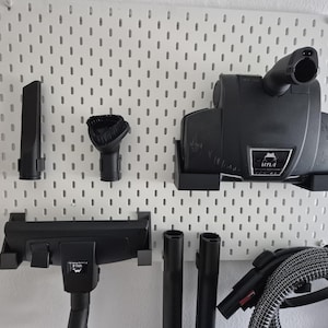 Puede incluir: Un tablero de clavijas blanco con varios accesorios de aspiradora, que incluyen una herramienta para grietas, un cepillo, una boquilla para el piso y una manguera.